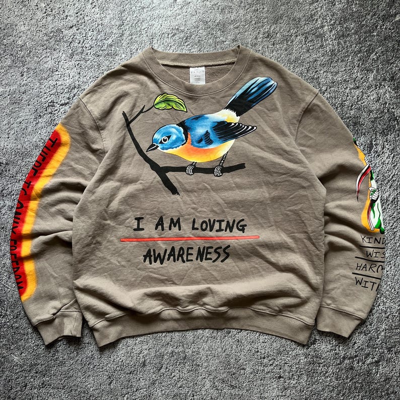 May include: A tan sweatshirt featuring a colorful bird perched on a branch. The bird has blue, yellow, and orange feathers. The text "I AM LOVING AWARENESS" is below the bird. The sleeves have additional text and graphics.