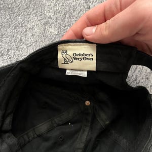 Puede incluir: Gorra de b&eacute;isbol negra con el logotipo y el texto de October's Very Own en una etiqueta. La etiqueta presenta un gr&aacute;fico de b&uacute;ho y las palabras "October's Very Own". La gorra est&aacute; hecha de tela oscura y tiene un bot&oacute;n en la parte superior.