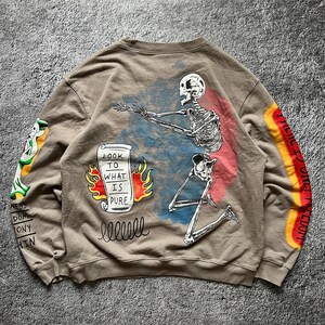 May include: A tan sweatshirt with a skeleton graphic on the back. The skeleton is kneeling with arms outstretched, with a scroll that reads "LOOK TO WHAT IS PURE" and flames. The sleeves have additional graphics.