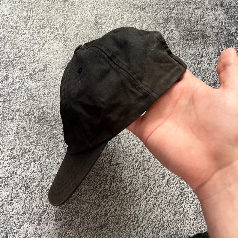 Puede incluir: Una gorra de b&eacute;isbol negra con visera curva. La gorra est&aacute; hecha de una tela oscura y tiene un dise&ntilde;o cl&aacute;sico de seis paneles. La gorra se sostiene en una mano sobre un fondo gris texturizado.