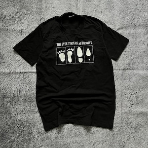 Puede incluir: Camiseta negra con el texto "THE EVOLUTION OF AUTHORITY" sobre un gráfico de huellas. El gráfico muestra una progresión de huellas, desde una pata de oso hasta una huella humana y, finalmente, un punto.