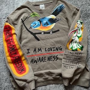 May include: A tan sweatshirt with a colorful bird perched on a branch. The left sleeve has the text "THERE IS ONLY FREEDOM" in red and yellow. The right sleeve features a Grim Reaper illustration and the words "KINDNESS, WISDOM, HARMONY, WITHIN". Below the bird are the words "I AM LOVING AWARENESS".