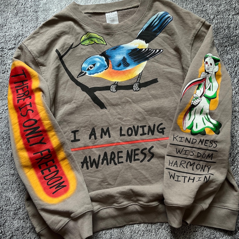 May include: A tan sweatshirt with a colorful bird perched on a branch. The left sleeve has the text "THERE IS ONLY FREEDOM" in red and yellow. The right sleeve features a Grim Reaper illustration and the words "KINDNESS, WISDOM, HARMONY, WITHIN". Below the bird are the words "I AM LOVING AWARENESS".