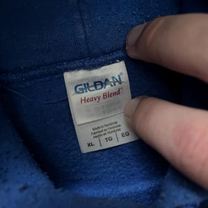 May include: Close-up of a blue hoodie with a white tag. The tag reads "GILDAN Heavy Blend" and includes size labels XL, TG, and EG. The tag also indicates the item was made in Honduras.