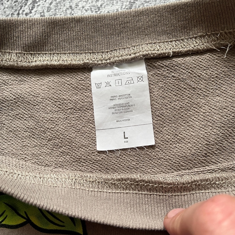 May include: Close-up of a tan sweatshirt with a white tag. The tag displays washing instructions, fabric composition (85% cotton, 15% polyester), and the size, which is L. The sweatshirt is made in China.