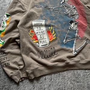 May include: A brown sweatshirt with a graphic design featuring a skeleton, a scroll with the text "LOOK TO WHAT IS PURE", and other illustrations. The sweatshirt has a crew neck and long sleeves.
