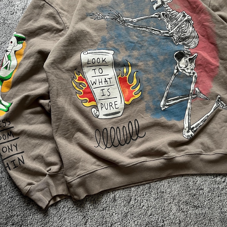 May include: A brown sweatshirt with a graphic design featuring a skeleton, a scroll with the text "LOOK TO WHAT IS PURE", and other illustrations. The sweatshirt has a crew neck and long sleeves.