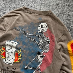May include: A brown sweatshirt featuring a skeleton graphic with outstretched arms. The design includes a scroll with the text "LOOK TO WHAT IS PURE" and flames. The skeleton is set against a blue and red background.
