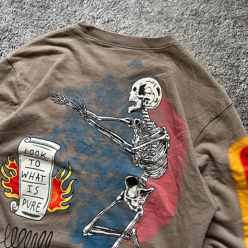 May include: A brown sweatshirt featuring a skeleton graphic with outstretched arms. The design includes a scroll with the text "LOOK TO WHAT IS PURE" and flames. The skeleton is set against a blue and red background.