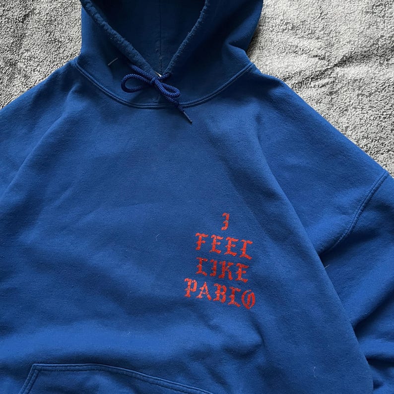May include: A blue hoodie with a drawstring and a front pocket. The hoodie features the text "I FEEL LIKE PABLO" in red, a design element. The garment is made of a soft material.