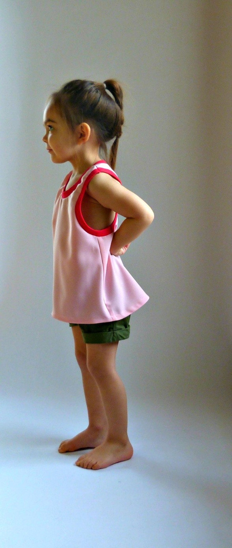 Puede incluir: Una ni&ntilde;a peque&ntilde;a con un vestido rosa sin mangas con ribete rojo y pantalones cortos verdes. Est&aacute; de pie sobre un fondo blanco.