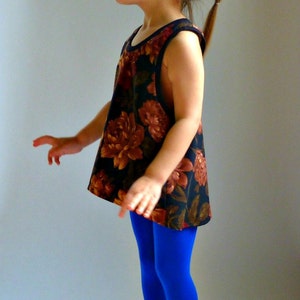 Puede incluir: Una ni&ntilde;a peque&ntilde;a con un top sin mangas de estampado floral y leggings azules. Est&aacute; de pie sobre una superficie blanca y mirando hacia un lado.