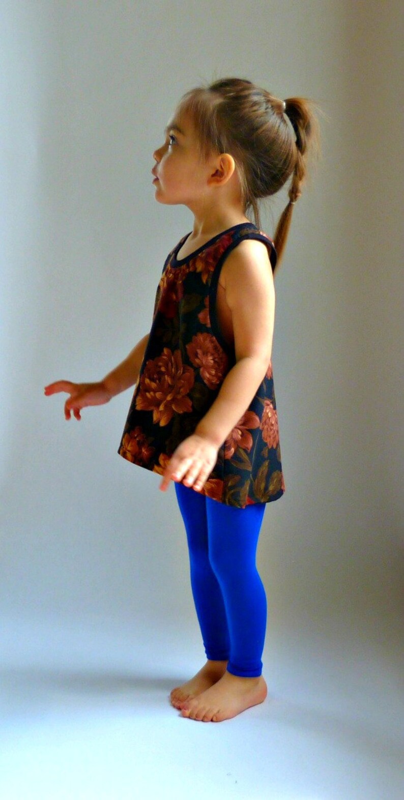 Puede incluir: Una ni&ntilde;a peque&ntilde;a con un top sin mangas de estampado floral y leggings azules. Est&aacute; de pie sobre una superficie blanca y mirando hacia un lado.