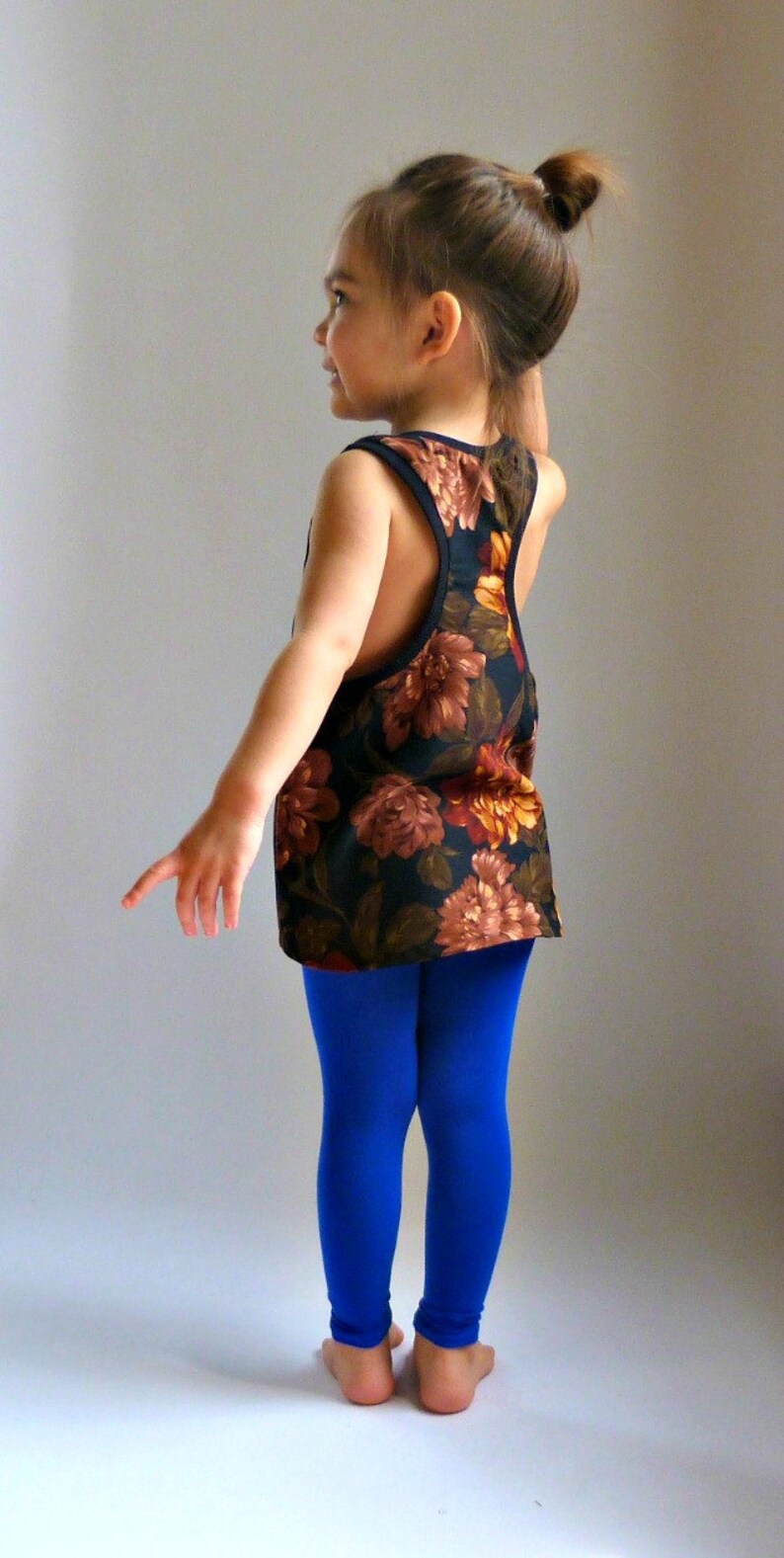 Puede incluir: Una persona joven lleva una camiseta sin mangas de estampado floral sobre fondo negro y leggings azules. La camiseta tiene un escote redondo y un estilo de espalda nadadora.