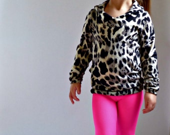Puede incluir: Una niña pequeña con una blusa de estampado de leopardo y leggings de color rosa brillante. Está de pie sobre un fondo blanco y mirando hacia un lado.