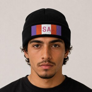 Puede incluir: Un gorro de punto acanalado negro con un parche rectangular en el dobladillo frontal. El parche muestra un diseño de bloques de color horizontales en morado, naranja y blanco, con las letras SA impresas en rosa oscuro en el centro de la sección blanca.