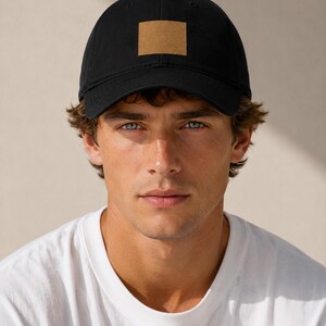 May include: A black baseball cap featuring a rectangular tan patch centered on the front panel. The cap has a structured crown, a curved brim, and a small button at the top center. The design is simple, with a solid black fabric base.