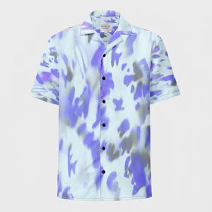 May include: Short-sleeved button-down shirt with a light blue base and an abstract pattern in shades of purple and gray. The shirt has a classic collar and black buttons. The text "ARTISTES CLOSET" is visible on the collar.