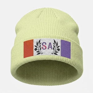 May include: A pale green beanie hat with a rectangular patch. The patch has the letters "SA" in a gradient colour, framed by black leaves and flowers, with red and purple stripes on either side.