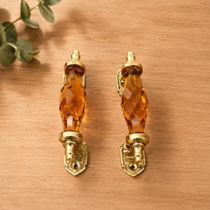 May include: Two cabinet handles featuring faceted amber-colored glass centers and polished brass metal ends. Each handle has a decorative brass mounting plate at the top and bottom with visible screw holes for attachment to furniture or cabinetry.