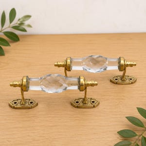 Può includere: Due maniglie per cassetti con una sezione centrale in vetro trasparente sfaccettato a forma di diamante. Ogni maniglia presenta finiture in metallo color ottone anticato con piastre di montaggio rettangolari decorate alle estremità con due fori.