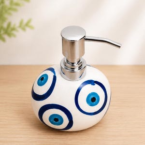 May include: A white ceramic soap dispenser with a rounded, bulbous shape featuring hand-painted blue and black evil eye motifs. The dispenser is topped with a shiny, metallic silver-toned pump mechanism designed for dispensing liquid soap.