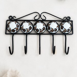 May include: A black metal wall-mounted rack featuring five hooks and five circular ceramic knobs. Each knob is white with a hand-painted brown floral vine pattern. The rack has a decorative scrolled top frame and a rectangular structure.