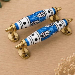 Op de afbeelding: Twee cilindervormige keramische deurklinken met een blauw en wit marmerpatroon, zwarte stippen en rode bladmotieven. De klinken zijn gemonteerd op messingkleurig metalen beslag met ovale voetplaten en puntige uiteinden aan beide zijden.