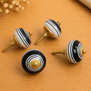 Puede incluir: Un juego de cuatro pomos redondos para armarios con un patrón de rayas blancas y negras. Cada pomo tiene una base de metal dorado pulido y un acento decorativo central a juego. Los pomos incluyen un perno roscado para su instalación.