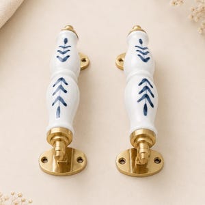 May include: Two vertical door handles featuring white ceramic bodies with hand-painted blue leaf patterns. Each handle is accented with polished brass hardware, including a rounded mounting plate with two screw holes at the base and a matching top cap.