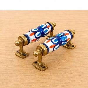 May include: Two drawer pulls featuring cylindrical ceramic handles with a white base, decorated with blue and orange painted patterns. The handles are mounted on antique brass-toned metal brackets with two screw holes on each base plate.