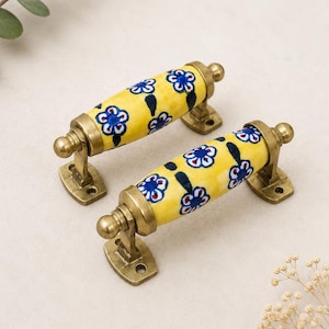 May include: Two cylindrical cabinet handles featuring a bright yellow ceramic surface with a repeating pattern of blue and white flowers and dark green leaves, secured by antique brass-toned metal brackets with circular mounting plates.