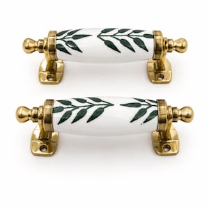 Può includere: Due maniglie per cassetti con un centro in ceramica bianca e un motivo a foglie verde scuro. Ogni maniglia presenta estremità e piastre di montaggio in ottone lucido con due fori per le viti, progettate per il fissaggio orizzontale.