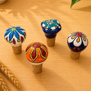 Può includere: Quattro tappi decorativi per bottiglie con teste in ceramica caratterizzate da motivi floreali e geometrici dipinti a mano in colori vivaci tra cui blu, arancione, verde e bianco, ciascuno fissato su una base in sughero naturale.