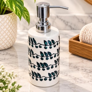 May include: A cylindrical ceramic soap dispenser with a glossy white finish and a repeating pattern of dark green vine and leaf motifs. The dispenser features a polished silver-tone metal pump mechanism at the top for dispensing liquid soap.