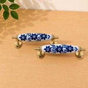May include: Two drawer pulls featuring white ceramic handles with a blue floral pattern. Each handle is mounted on gold-toned metal hardware with two oval-shaped base plates, each containing two screw holes for secure attachment to furniture.