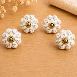 May include: Four white ceramic drawer knobs with a scalloped, flower-like shape. Each knob features a central metallic gold-toned circular accent and delicate gold-painted leaf patterns radiating outward from the centre to the scalloped edges.
