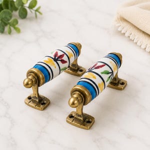 Op de afbeelding: Twee cilindervormige keramische ladeknoppen met een witte basis en bloemmotieven in blauw, geel en rood. Elke handgreep is gemonteerd op een metalen voet met een antieke messing afwerking en twee schroefgaten voor bevestiging aan meubels.
