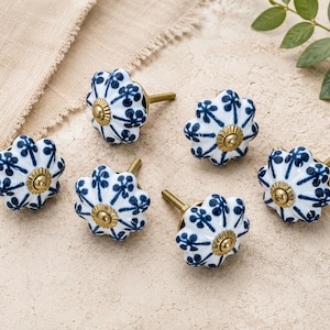 Peut inclure: Un ensemble de six boutons de tiroir ronds en céramique présentant une base blanche avec un motif floral bleu et un composant central en métal doré. Chaque bouton a une forme festonnée rappelant une citrouille avec une finition lisse.