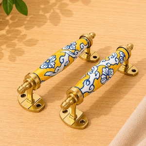 Op de afbeelding: Twee kastgrepen met een cilindrisch geel keramisch middenstuk met een blauw en wit bloemmotief. Elke greep wordt ondersteund door twee messingkleurige metalen montageplaten met schroefgaten, verbonden door bijpassend messingkleurig beslag.
