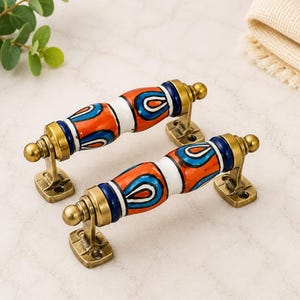 May include: Two drawer pulls featuring a cylindrical ceramic handle with a white, orange, and blue teardrop pattern. The handles are mounted on antique brass-toned metal brackets with two screw holes each, finished with small spherical end caps.