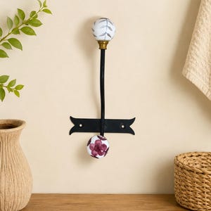 May include: A black metal wall hook featuring two round ceramic knobs. The top knob is white with a grey leaf pattern, while the bottom knob is white with a dark red floral design. The hook is mounted on a horizontal black metal plate.