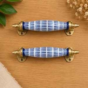 May include: Two drawer pulls featuring a cylindrical ceramic center with a white and blue vertical striped pattern. Each pull is secured by brass-toned metal hardware at both ends, which includes mounting plates with screw holes for installation.