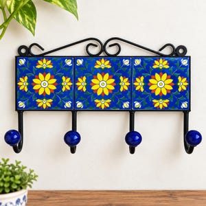 Puede incluir: Un perchero de pared con marco de metal negro y volutas decorativas en la parte superior. Incluye tres azulejos de cerámica cuadrados con fondo azul y un patrón floral amarillo, y cuatro ganchos negros con pomos de cerámica azul.