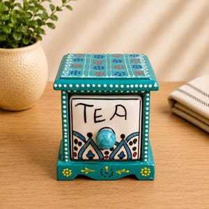 May include: A square ceramic tea storage box with a teal frame and a white drawer front labeled with the word TEA in black script. The top features a teal grid pattern with red and blue dots, while the base displays yellow floral accents.