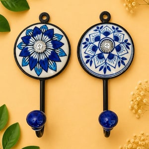 May include: Two wall hooks featuring circular ceramic plates with blue and white floral patterns. Each hook has a black metal frame, a central silver-toned screw, and a small blue spherical knob at the bottom of the hook arm for hanging items.