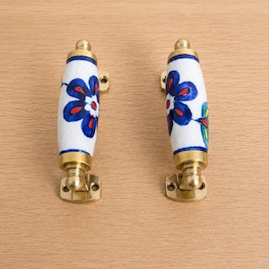 May include: Two cylindrical ceramic door handles featuring a white base with hand-painted blue flowers, red centres, and green leaf accents. Each handle is secured by polished brass metal hardware at both ends, including mounting plates.