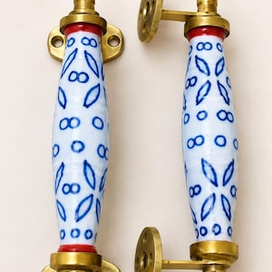 Puede incluir: Dos tiradores de puerta decorativos. Cada tirador tiene un cuerpo de cerámica blanca con un diseño floral azul, detalles rojos y herrajes de latón. Los tiradores están diseñados para una fácil instalación y añaden elegancia.