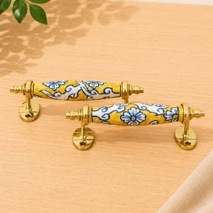 May include: Two drawer pulls featuring a cylindrical ceramic centre with a yellow background and blue floral patterns. The handles are supported by polished gold-tone metal hardware at each end, including oval mounting plates with screw holes.