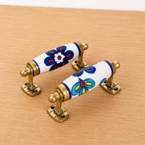 Op de afbeelding: Twee cilindrische keramische ladeknoppen met handgeschilderde bloemmotieven in blauw, rood en geel. Elke handgreep is gemonteerd op een metalen voet met een antieke messing afwerking en twee schroefgaten voor bevestiging aan een meubel.